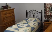 Small bedroom with a single bed covered by a patterned quilt, a wooden dresser and nightstand, and wall decorations.