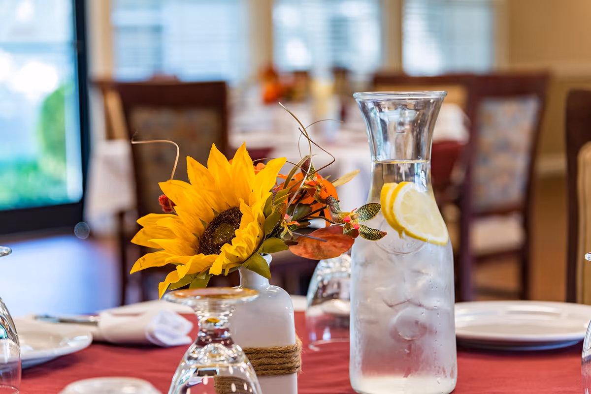 Close-up of a dining table set with a vase holding a bright yellow sunflower and autumn-themed decorations, a glass pitcher filled with ice water and lemon slices, and neatly arranged plates and napkins in a softly lit dining room.