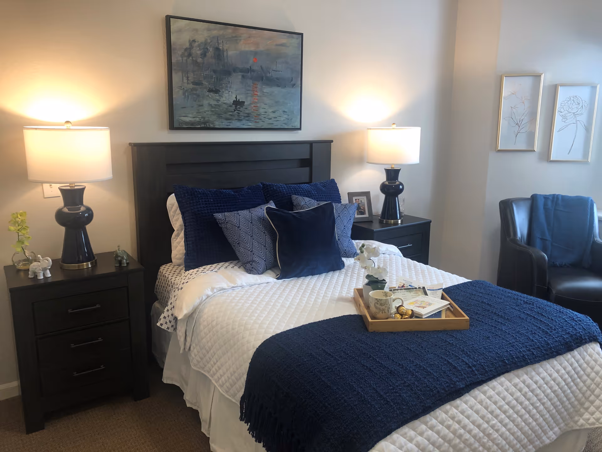 A neatly made bed with white and navy blue bedding, including multiple pillows and a navy blue throw blanket. Two dark wooden nightstands flank the bed, each with a lamp and decorative items. Above the bed hangs a framed painting. To the right, there is a dark leather armchair with a blue throw draped over it, and two framed floral line drawings on the wall.