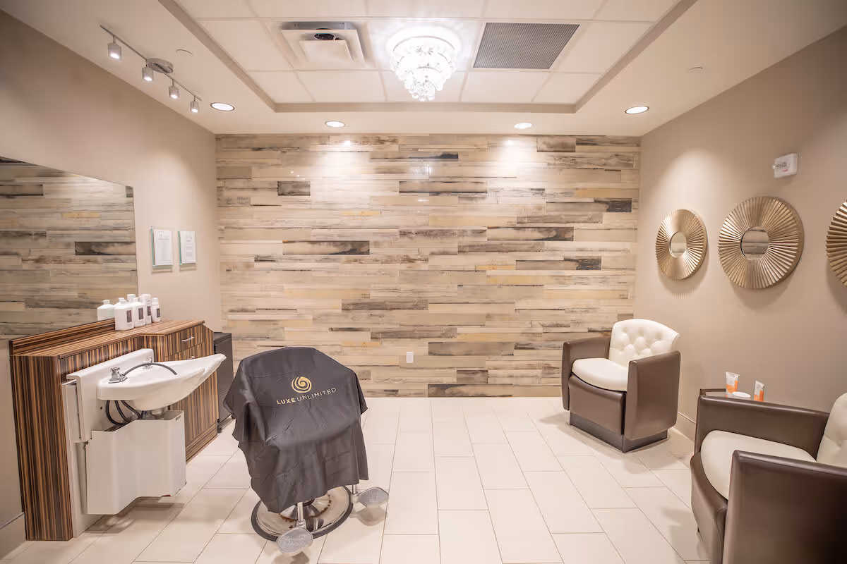 A modern salon area with a hair washing station on the left, a salon chair covered with a black Luxe Unlimited cape in the center, and two brown and white cushioned armchairs on the right. The back wall features a wood panel design with spotlights above, and three decorative round mirrors hang on the right wall. The floor is tiled in light beige, and the ceiling has recessed lighting and a small chandelier.