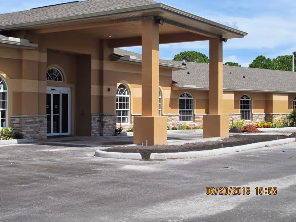 Exterior view of the Gardens of Venice senior living facility showing the entrance with a covered drop-off area, beige and tan walls with stone accents, arched windows, and a paved driveway under a partly cloudy sky.