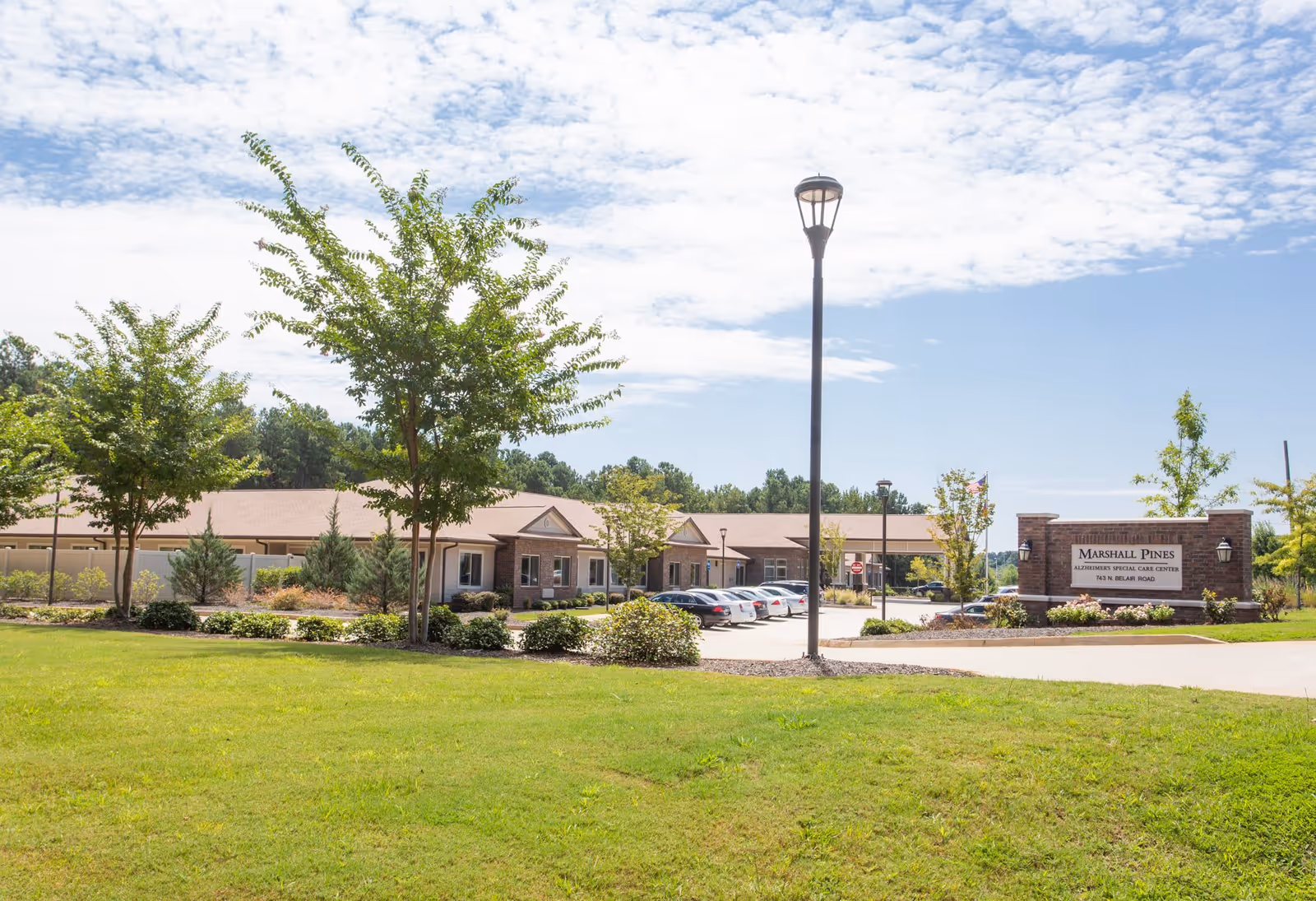Front exterior of Marshall Pines Assisted Living & Memory Care with a lawn, trees, parking lot, and entrance sign.
