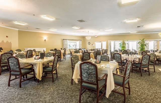 A spacious dining room with multiple tables covered in beige tablecloths, each surrounded by wooden chairs with green patterned cushions. The room has carpeted floors, large windows with curtains, and soft overhead lighting. There are plants near the windows and a clock on the wall.
