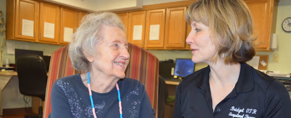 An elderly woman with gray hair wearing a blue sweater and colorful beaded necklace is smiling and looking at a woman with short blonde hair wearing a black polo shirt with the name Bridget OTR Occupational Therapist embroidered on it. They are seated in a room with wooden cabinets, a computer, and office chairs in the background.