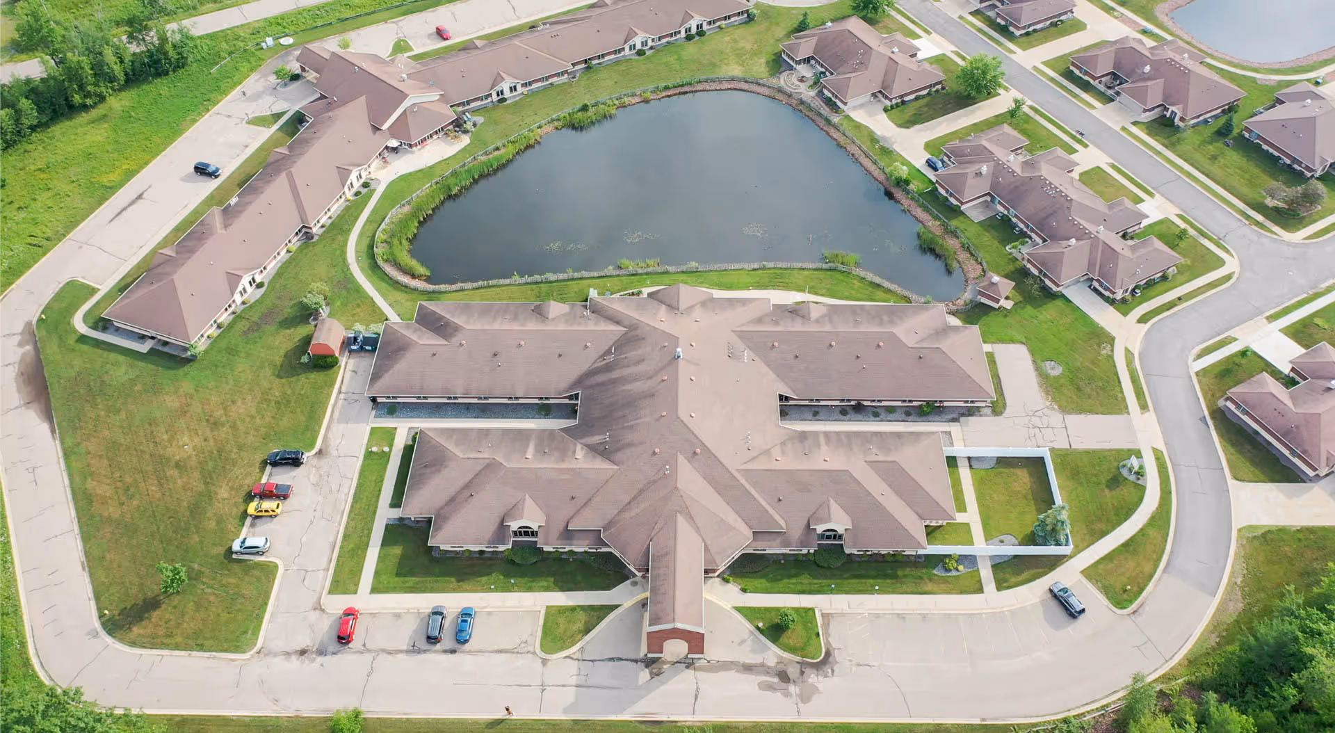 Aerial view of a senior living complex with a central building, surrounding cottages, parking areas, roads, and a pond.