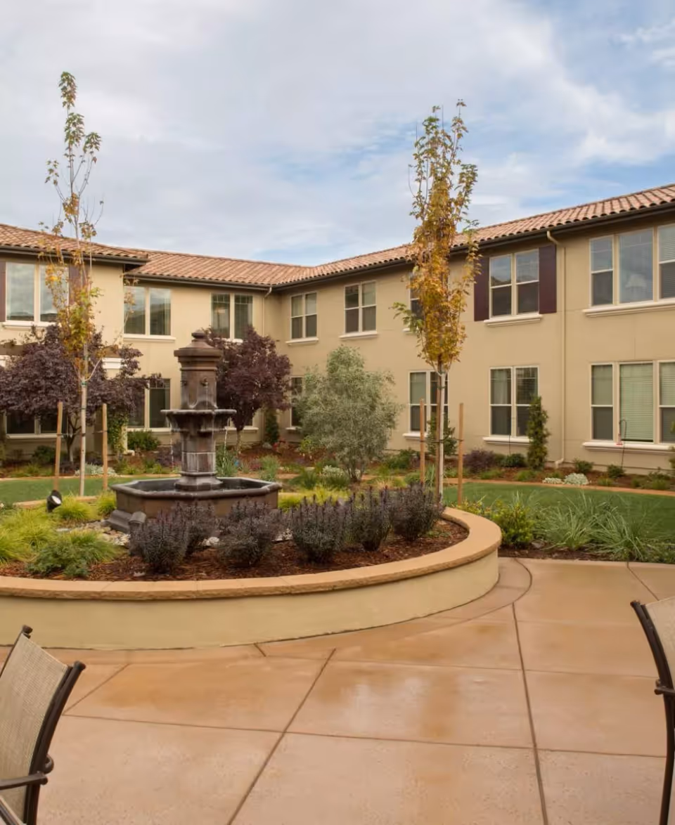 Outdoor courtyard area of a senior living facility with a central stone fountain surrounded by landscaped plants and young trees. The courtyard is paved with concrete and has seating chairs visible at the edges. The building surrounding the courtyard has beige walls, multiple windows, and a tiled roof under a cloudy sky.