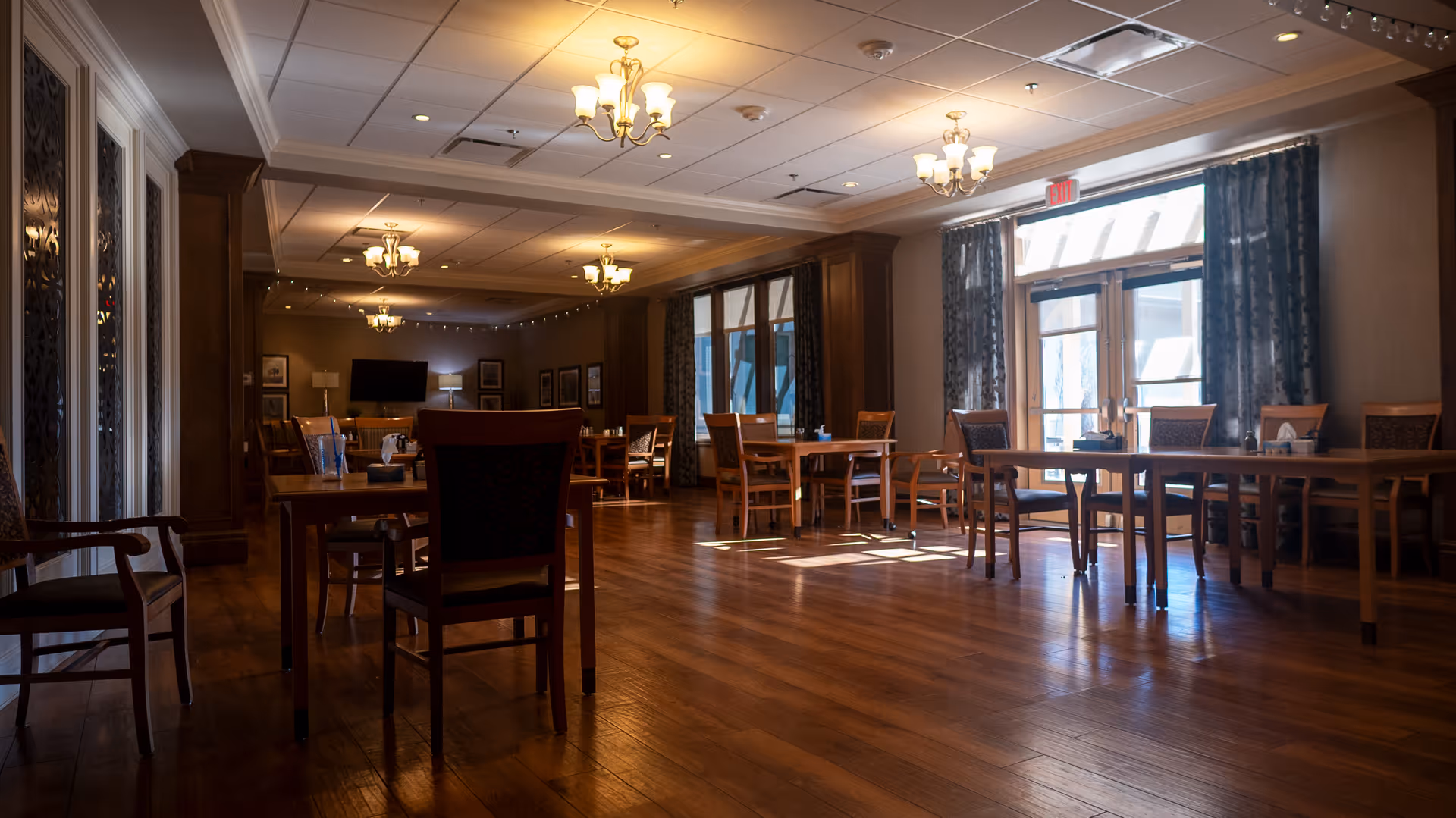 A spacious dining room with wooden floors and multiple wooden tables and chairs arranged neatly. The room is softly lit by several ceiling chandeliers and natural light coming through large windows with curtains. There is a television mounted on the far wall and framed pictures hanging on the walls.