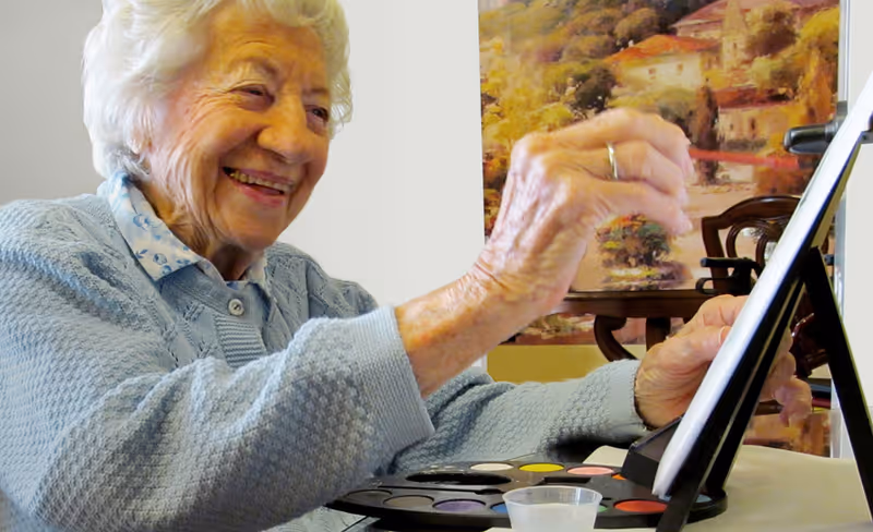 An elderly woman smiling and painting on a canvas with a paintbrush, sitting at a table with a palette of watercolors and a small cup of water. A colorful painting and a wooden chair are visible in the background.