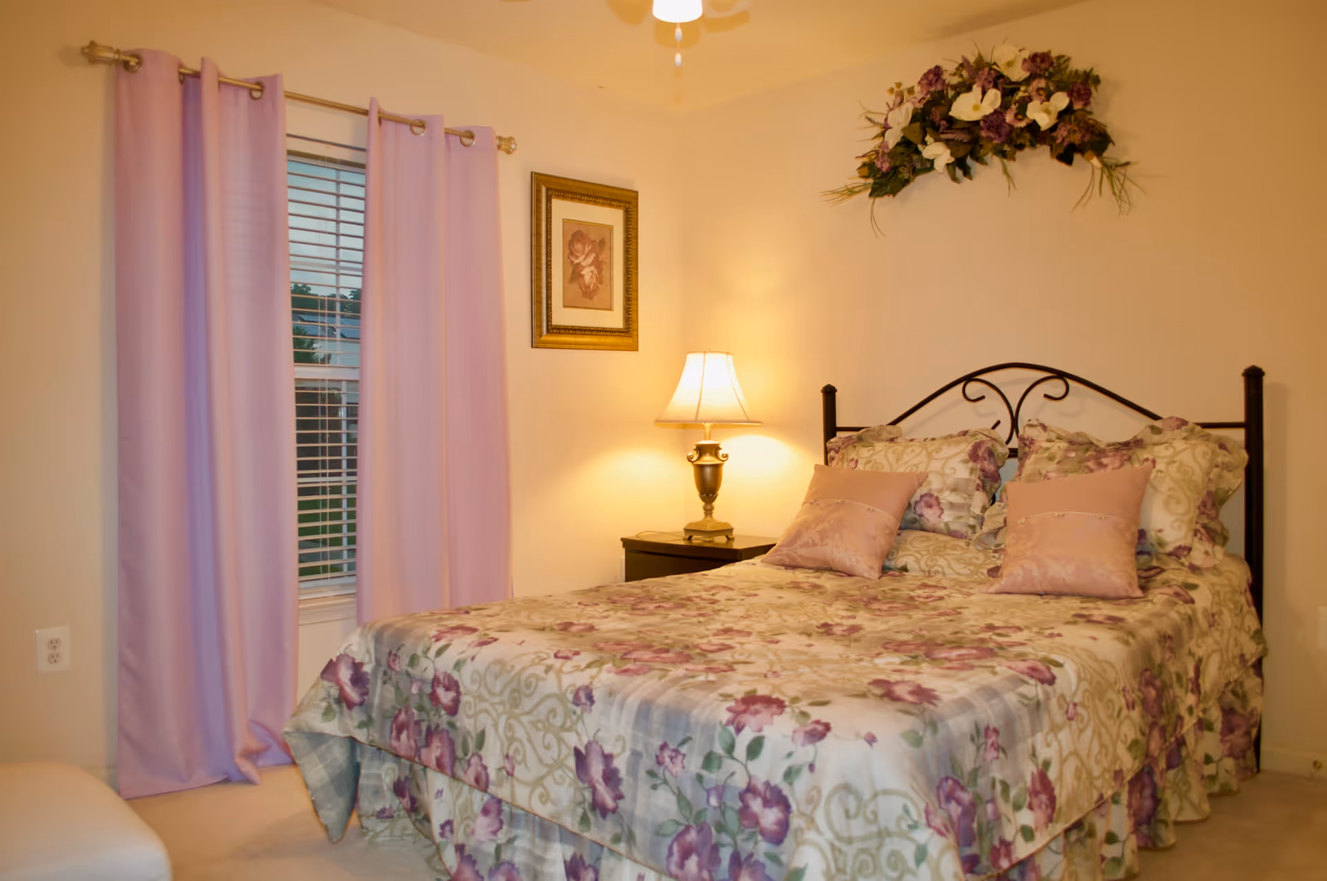 A cozy bedroom with a bed covered in floral-patterned bedding in shades of purple and cream. The bed has matching pillows and a black metal headboard. To the right of the bed is a nightstand with a decorative lamp emitting warm light. Above the bed is a floral wall decoration. On the left side of the room, there is a window with closed lavender curtains and white blinds. A framed picture hangs on the wall between the window and the lamp.