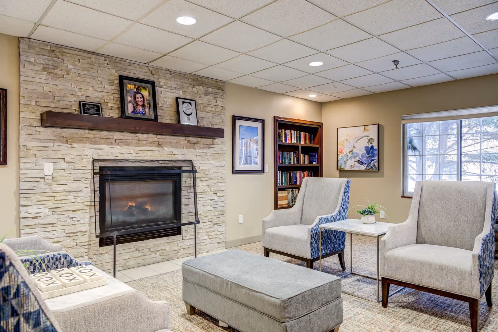 Cozy senior living room with a stone fireplace, two upholstered armchairs, a cushioned ottoman, a small side table with a plant, a bookshelf filled with books, framed artwork on the walls, and a large window letting in natural light.