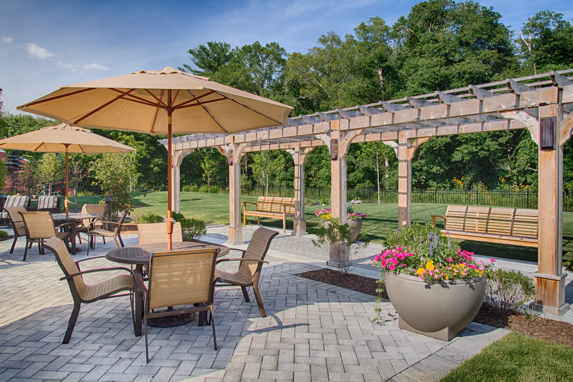 Sunlit outdoor patio with tables and umbrellas, a wooden pergola, benches, and flower planters on a paved area next to a lawn.