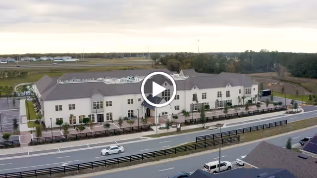 Aerial view of a large, two-story senior living facility building with a beige exterior and dark roof, surrounded by parking lots, roads, and some greenery in the background under a cloudy sky.