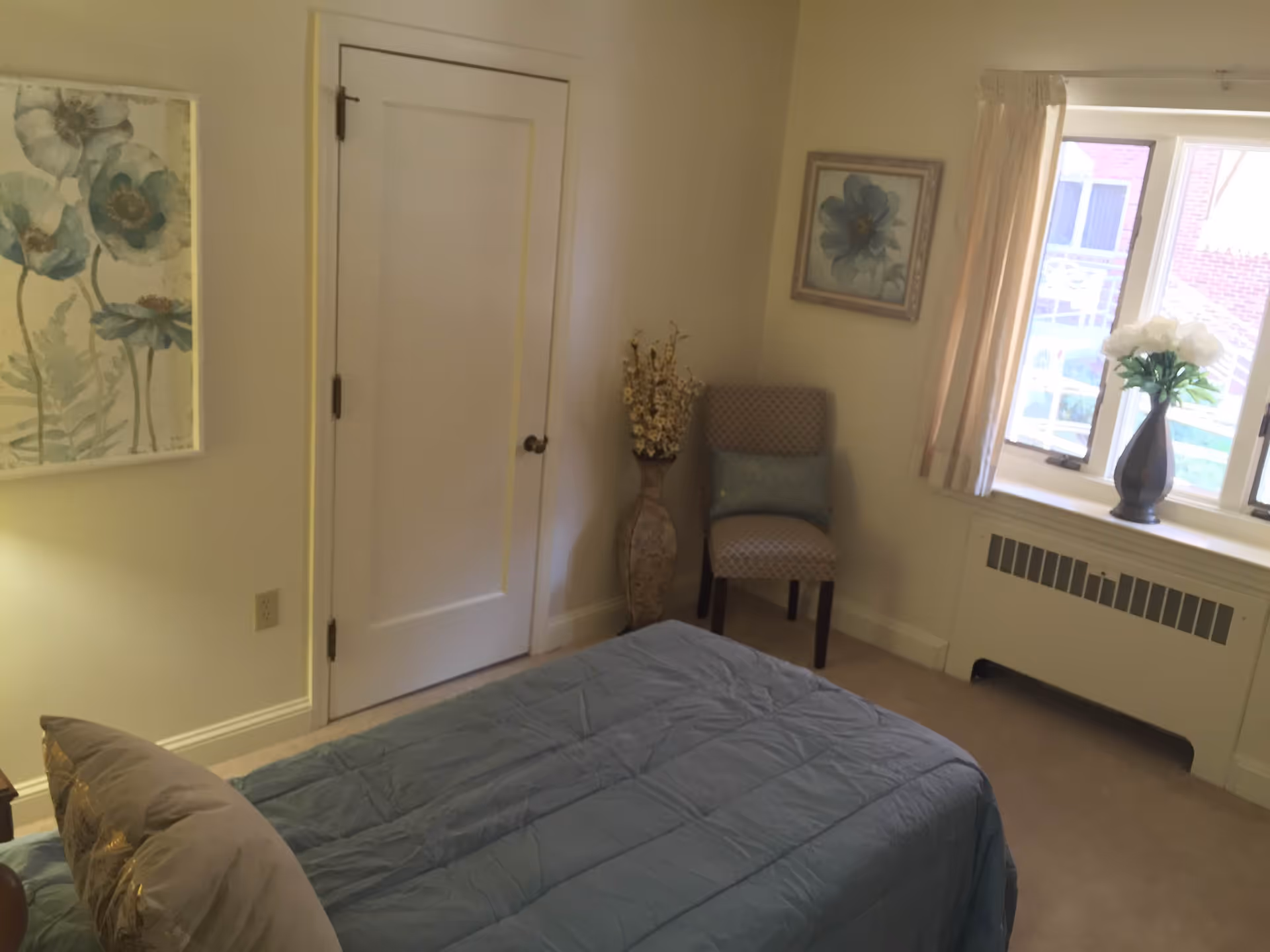 A bedroom with a bed covered in a blue comforter and a pillow. There is a closed white door on the left wall, a chair with a cushion in the corner, and a tall vase with dried flowers next to it. Two framed floral paintings hang on the walls. A window with cream-colored curtains lets in natural light, and a vase with white flowers sits on the windowsill above a radiator.