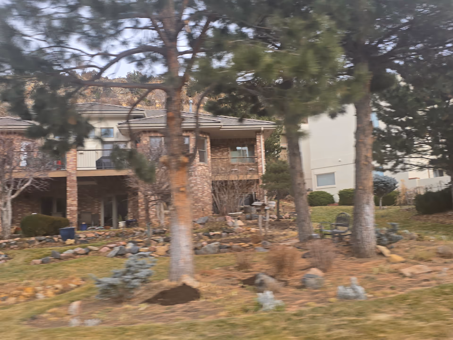 View of a brick building with a covered patio area, surrounded by trees, rocks, and landscaped garden beds with shrubs and grass.