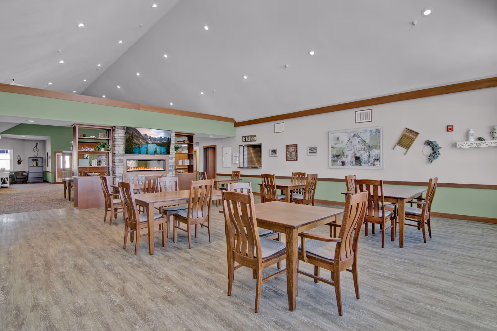 A spacious dining area in a senior living facility with several wooden tables and chairs arranged neatly. The room has a high vaulted ceiling with recessed lighting, light wood flooring, and walls painted in a combination of light green and white. There is a stone fireplace with a TV mounted above it, and shelves with decorative items on either side. The room has a cozy and welcoming atmosphere.