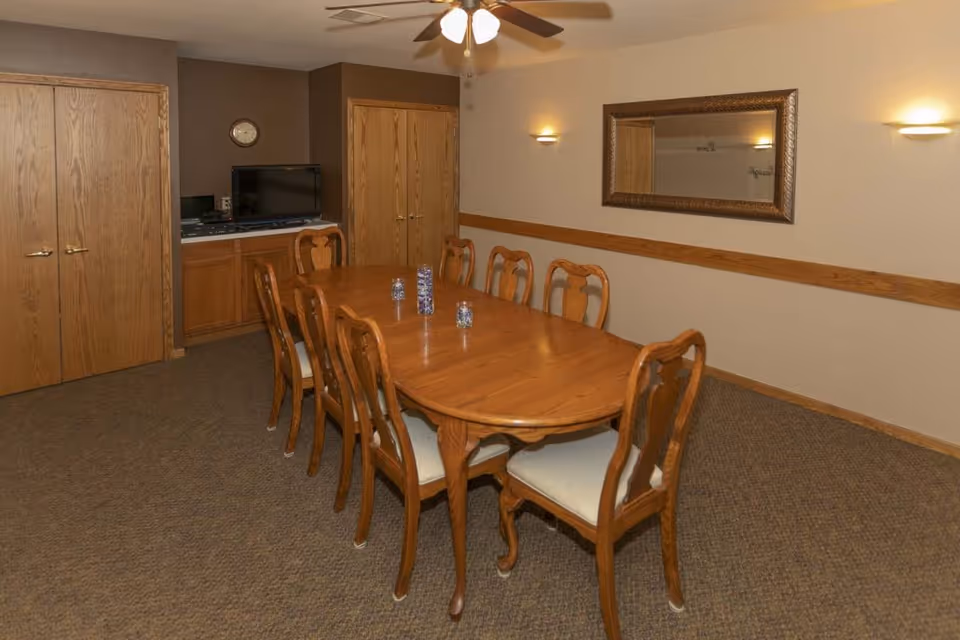 A dining room with a long wooden table surrounded by eight wooden chairs with cushioned seats. The room has beige walls with a wooden chair rail, a large decorative mirror on one wall, two wall sconces, a ceiling fan with lights, and a carpeted floor. In the corner, there is a small counter with a TV and some cabinets beneath it, flanked by two wooden doors.