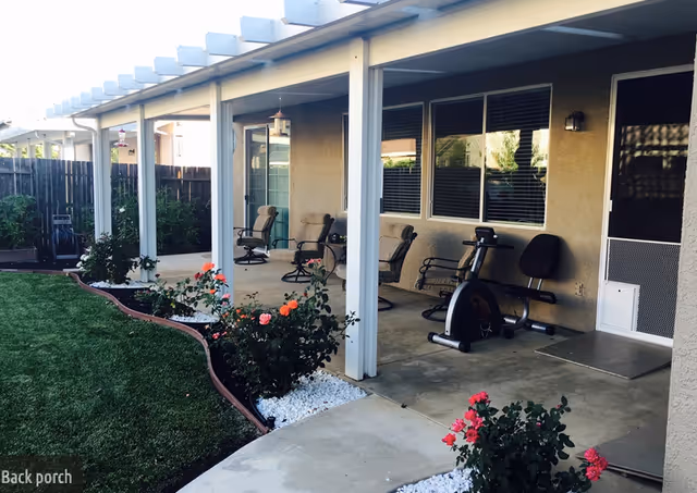 Covered back porch area with several cushioned chairs, a stationary exercise bike, and a garden with blooming rose bushes and green grass. The porch has white pillars and a concrete floor, with a wooden fence enclosing the garden area.