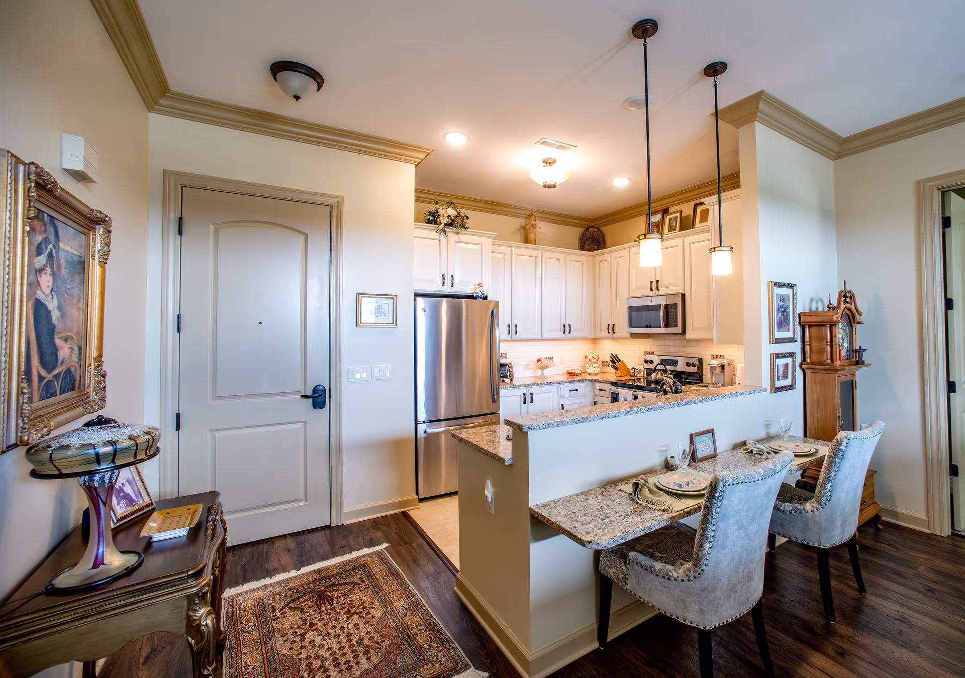 Open-plan kitchen with white cabinets, stainless steel refrigerator and a granite breakfast bar with two upholstered chairs and decorative furnishings.