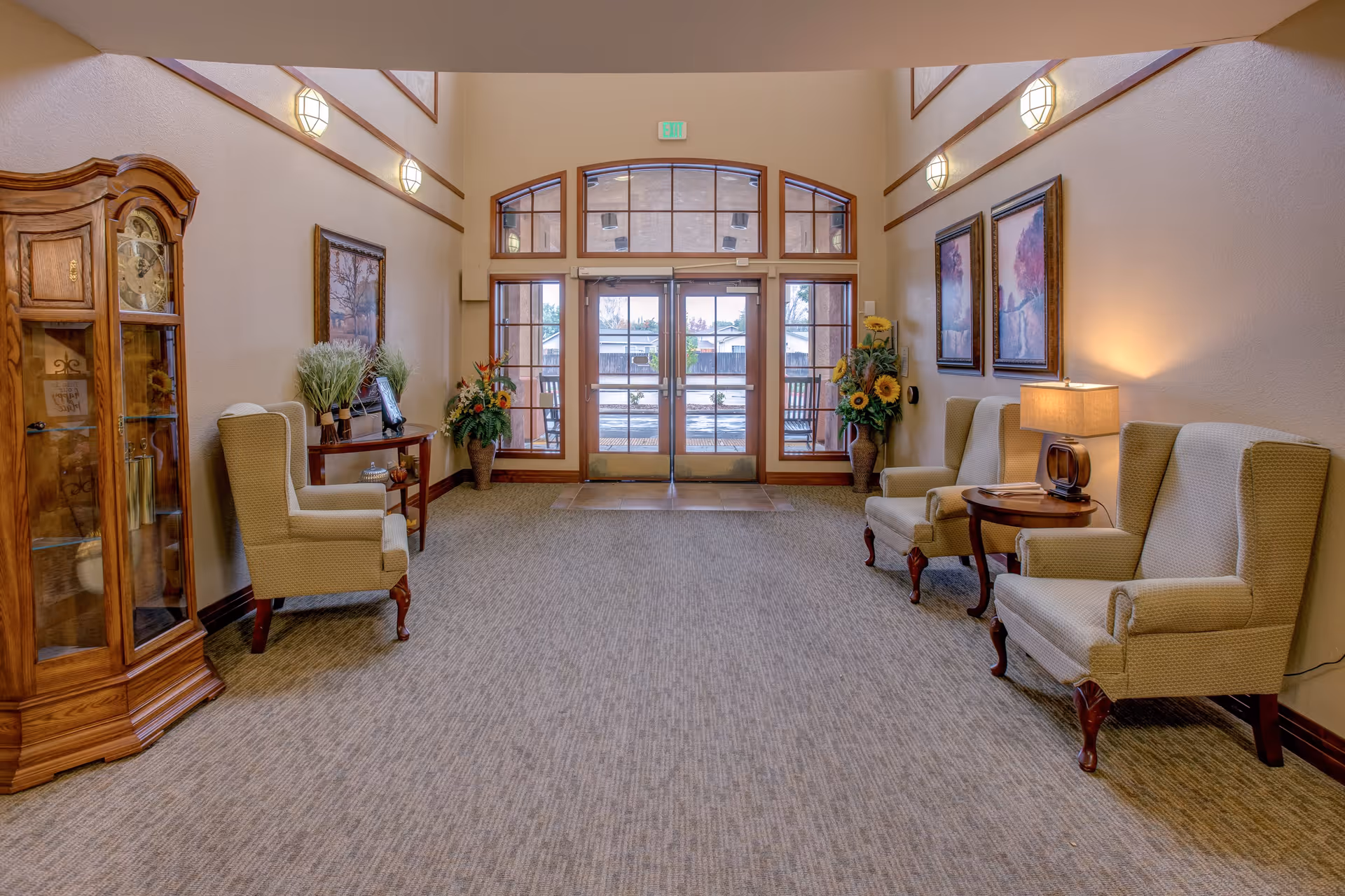 A bright and welcoming lobby area with beige walls and carpeted floor. The space features four upholstered armchairs arranged along the walls, a wooden grandfather clock on the left, a small wooden table with a lamp and decorative items on the right, and two framed paintings on the right wall. Large glass double doors with windows above and on the sides allow natural light to enter, and there are floral arrangements near the doors and on a small table on the left side.