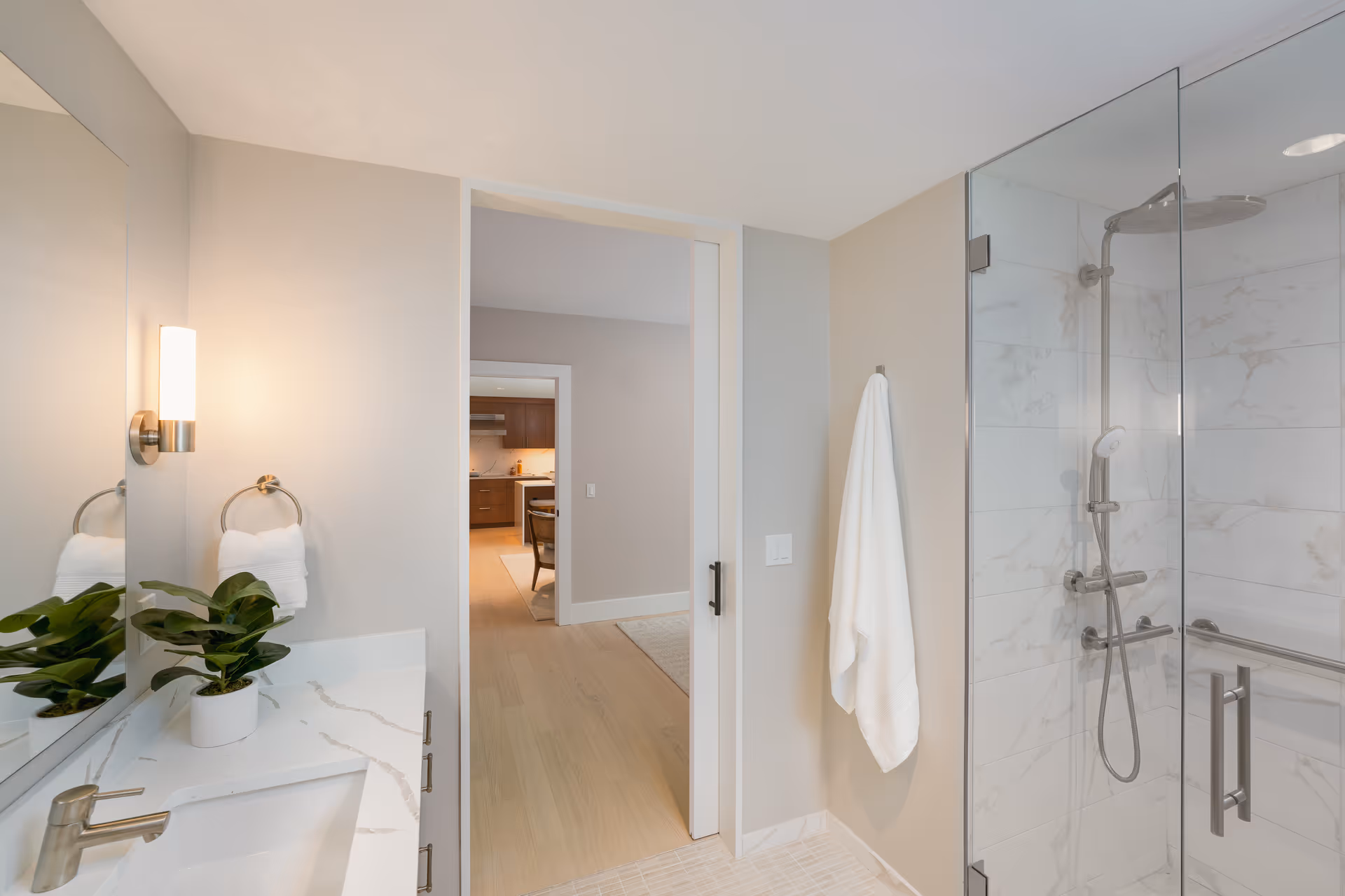 A modern bathroom featuring a glass-enclosed shower with a rainfall showerhead and handheld shower. The walls inside the shower are tiled with white marble-patterned tiles. A white towel hangs on a hook outside the shower. To the left, there is a white countertop with a sink, a silver faucet, a small potted plant, and a towel ring holding a white hand towel. The bathroom opens into a hallway leading to a kitchen area with wooden cabinets and a dining table with chairs.
