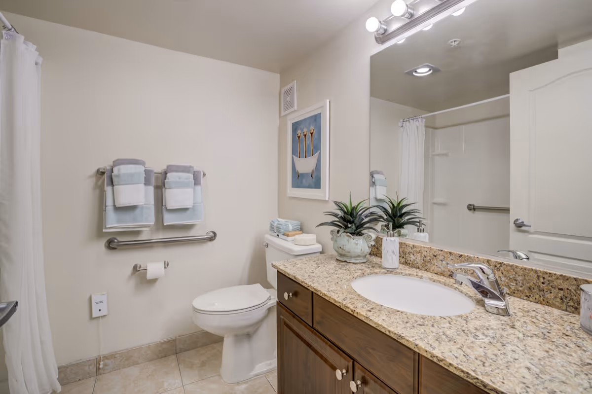 Clean, well-lit bathroom featuring a granite countertop vanity with sink and mirror, toilet, towel rack with folded towels, and a shower with curtain.