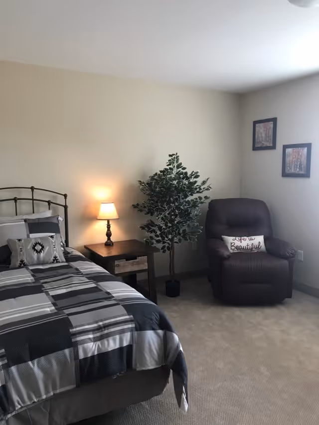 A bedroom with a bed covered in a gray and white checkered comforter, a wooden nightstand with a lit lamp, a potted artificial plant, and a dark brown recliner chair with a pillow that says 'Life is Beautiful'. Two framed pictures hang on the wall near a window.