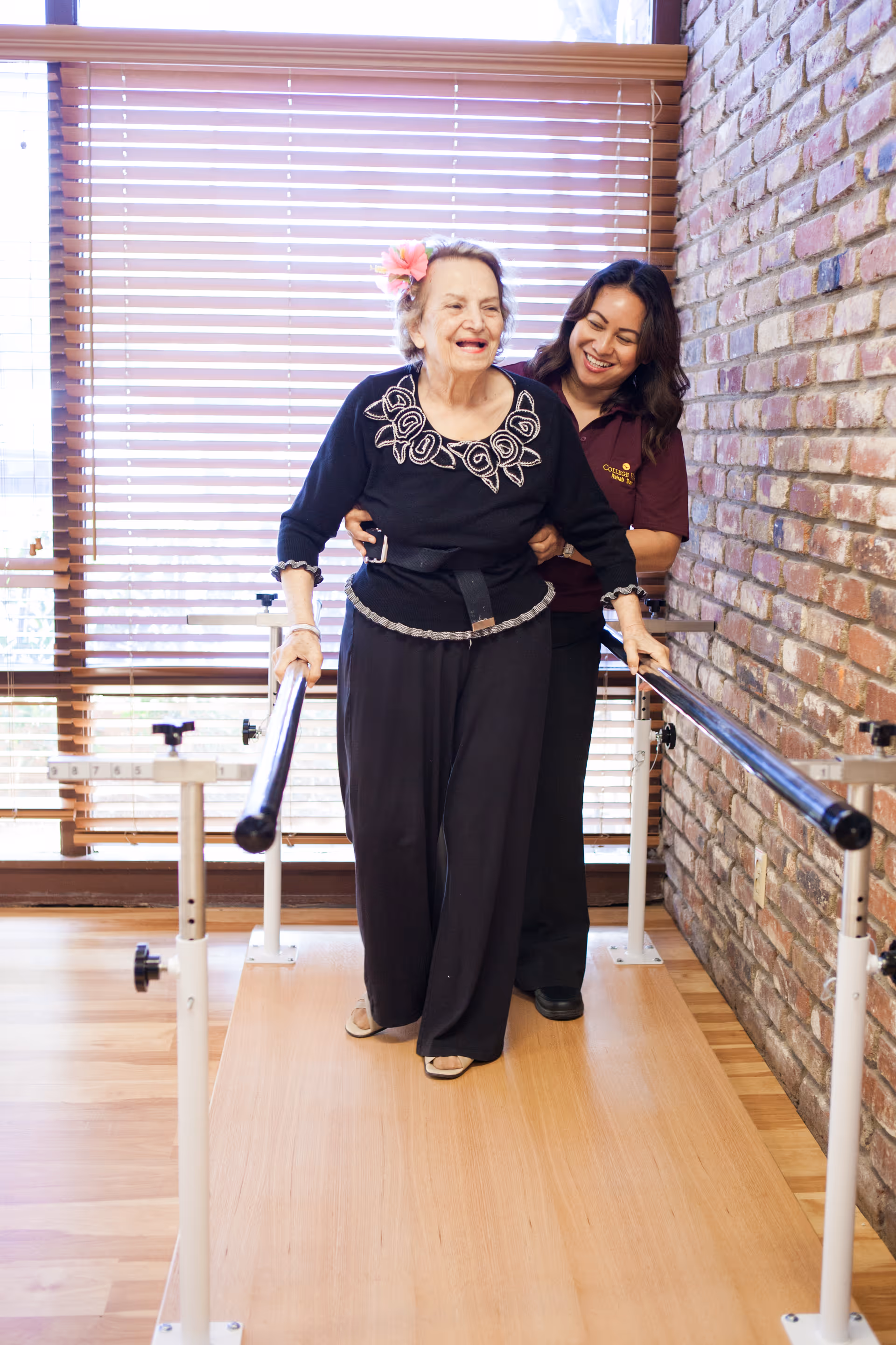 An elderly woman with a flower in her hair is walking with the support of parallel bars while being assisted by a smiling caregiver in a maroon shirt. They are indoors with wooden flooring, a brick wall on one side, and large windows with horizontal blinds in the background.