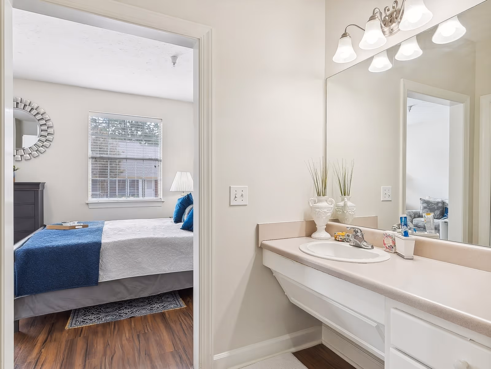 View of a bathroom sink and countertop with a large mirror and light fixtures above. Through an open doorway, a bedroom is visible with a bed covered in white and blue bedding, a window with blinds, a bedside lamp, and a decorative round mirror on the wall.