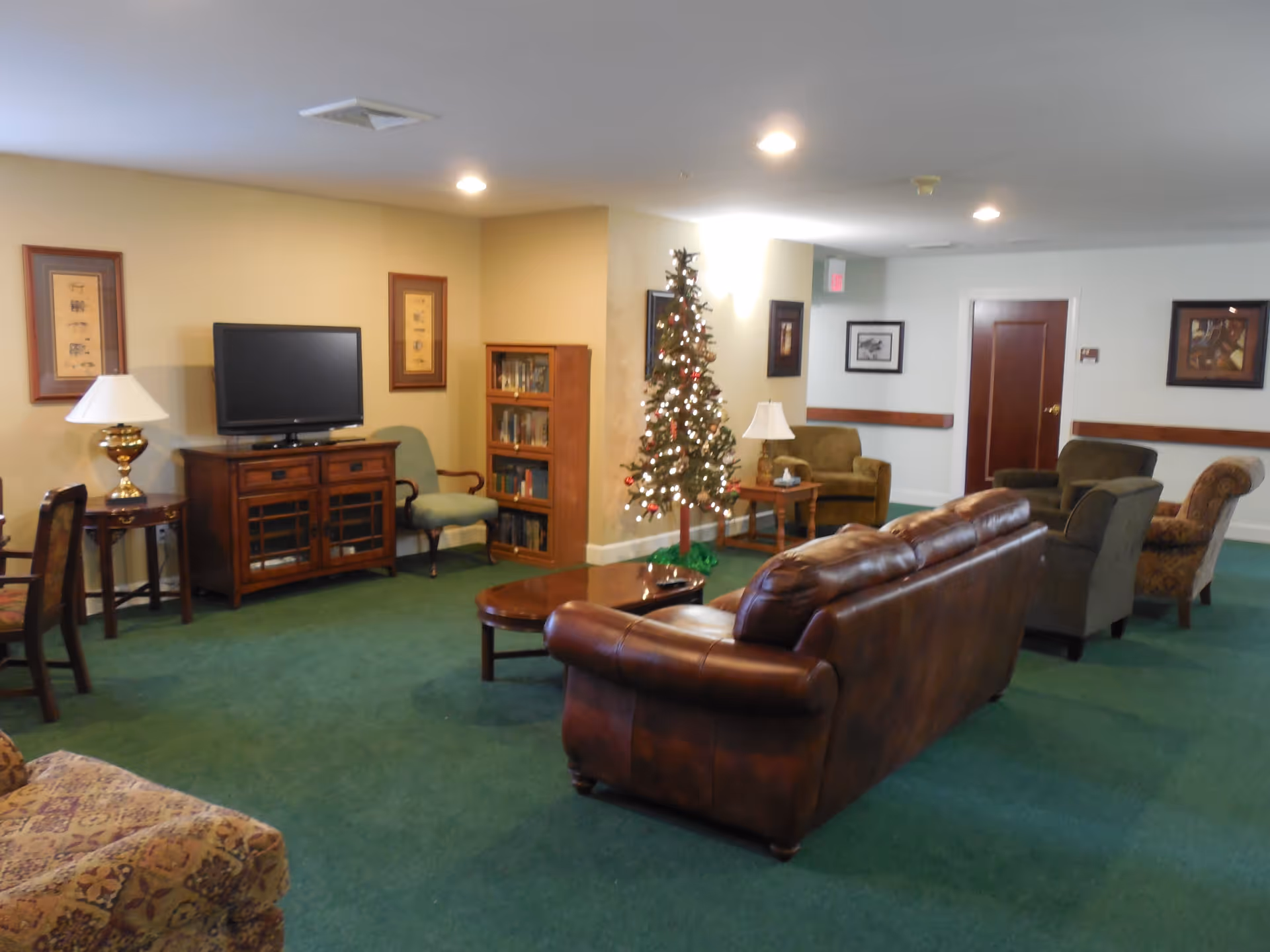 A cozy living room area with green carpet, a brown leather sofa, several upholstered chairs, a wooden coffee table, a TV on a wooden stand, a small bookshelf, framed pictures on the walls, and a decorated Christmas tree with lights.