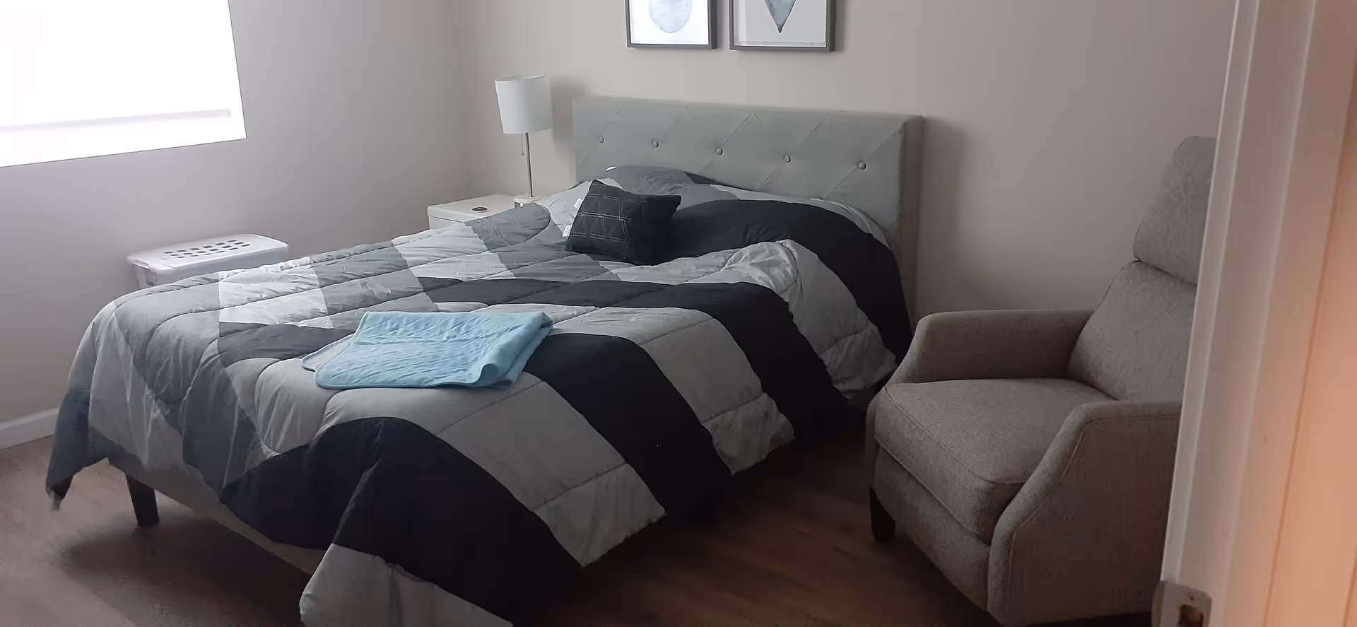 A bedroom with a bed covered in a gray and black geometric patterned comforter, a light blue towel folded on top, a small black pillow, a gray upholstered armchair, a white nightstand with a lamp, and a window letting in natural light.