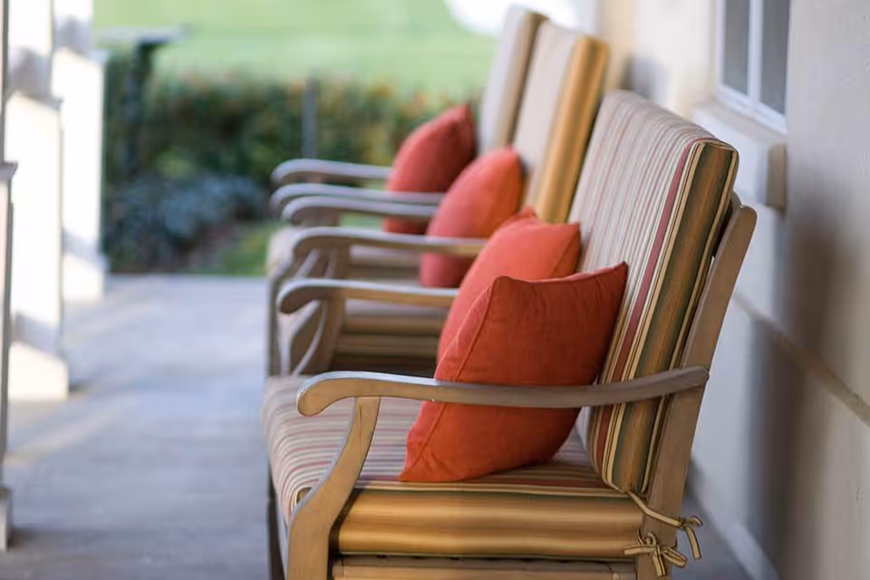 A row of outdoor cushioned chairs with striped upholstery and red pillows lined up along a porch or patio area with a blurred garden in the background.
