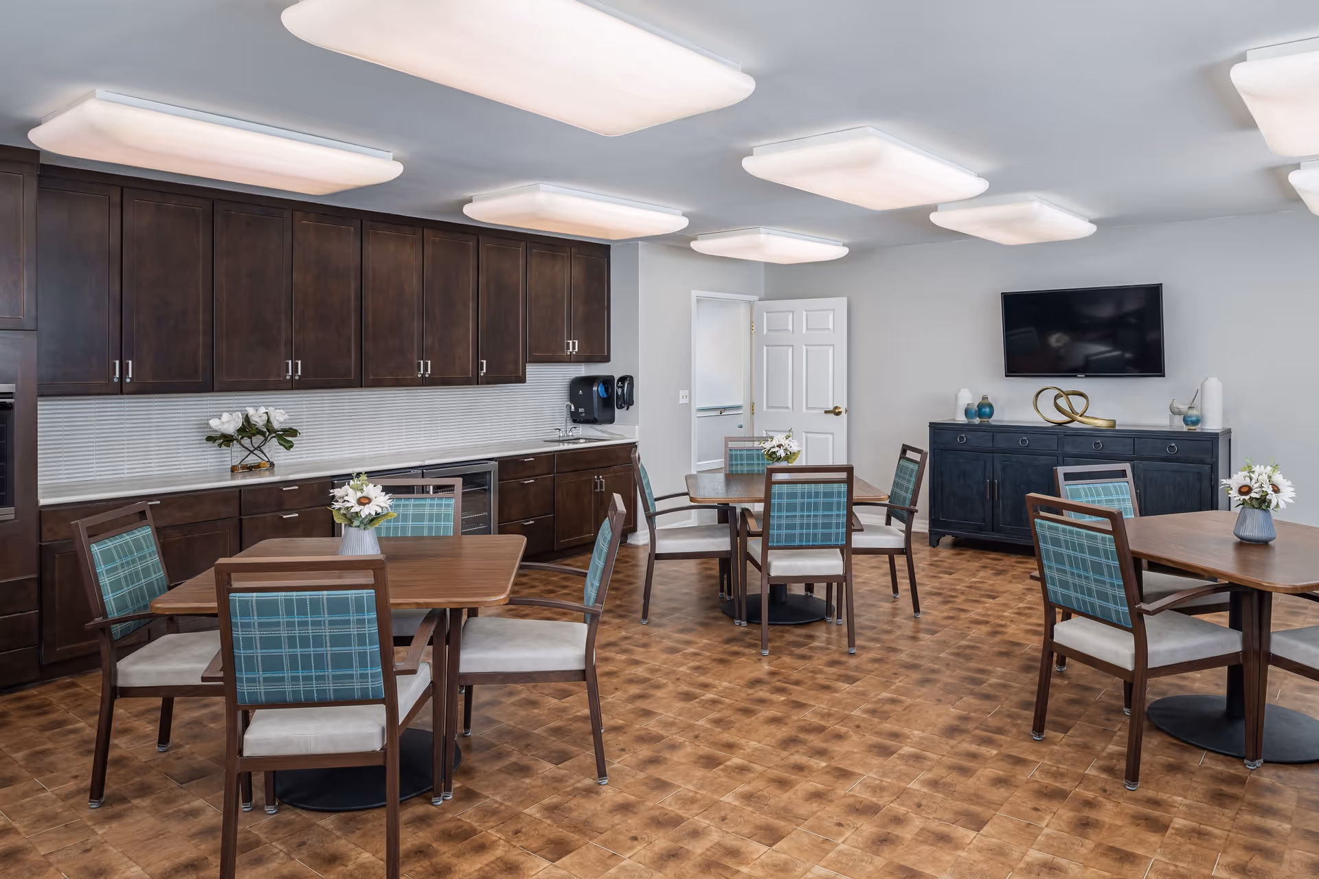 Bright dining/activity room with several wooden tables and plaid-upholstered chairs, a kitchenette with dark cabinets, and a wall-mounted TV.