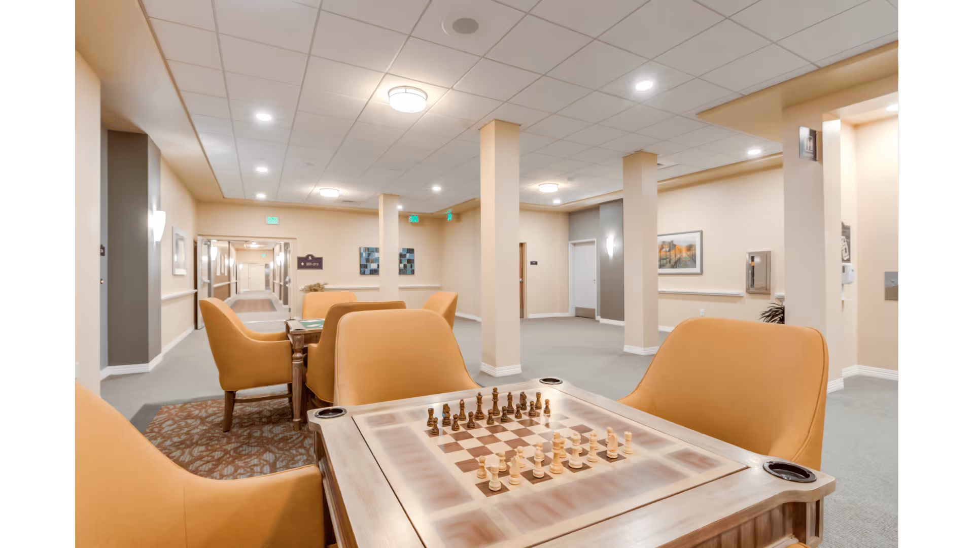 A well-lit common area in a senior living facility featuring a chess table with chess pieces set up and surrounded by comfortable tan armchairs. The room has beige walls, carpeted floors, and several columns. There are framed artworks on the walls and a hallway visible in the background.