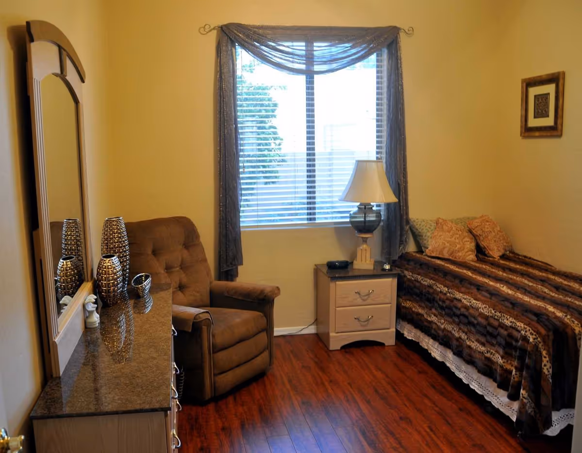 A cozy bedroom with a single bed covered in a brown patterned blanket and several pillows. Next to the bed is a small nightstand with a lamp and an alarm clock. A comfortable brown armchair is positioned near a window with blinds and sheer curtains. A dresser with a large mirror and decorative vases is on the left side of the room. The floor is wooden, and the walls are painted light yellow.