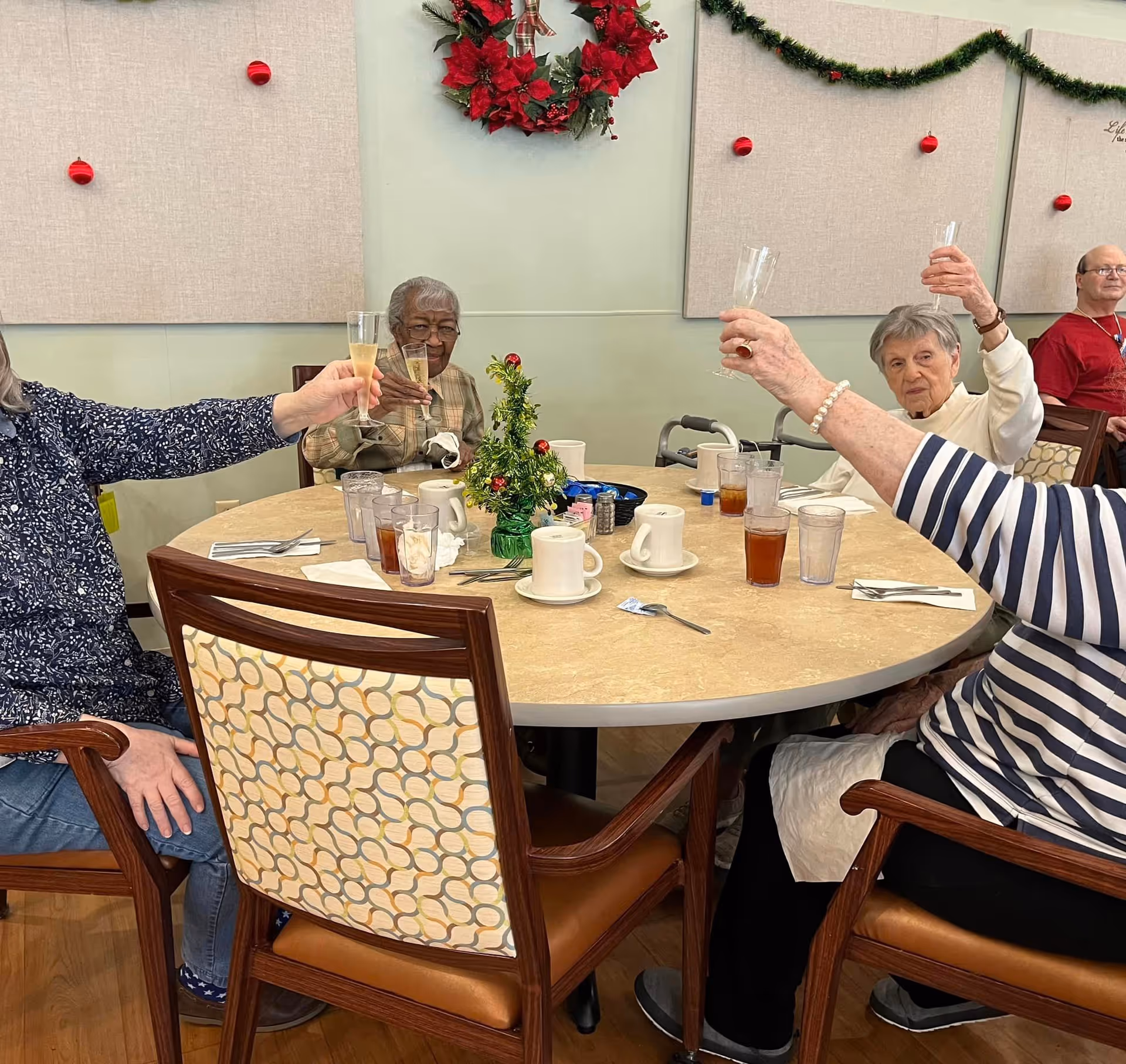 A group of elderly people sitting around a round dining table raising glasses in a toast. The table has cups, glasses with iced tea, utensils, and a small decorated Christmas tree centerpiece. The room is decorated with a red wreath and garland with red ornaments on the wall.