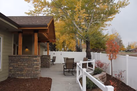 Outdoor patio area with stone pillars and wooden roof overhang attached to a building. There are several chairs and tables on a concrete walkway next to a white fence. Trees with autumn-colored leaves are visible in the background.