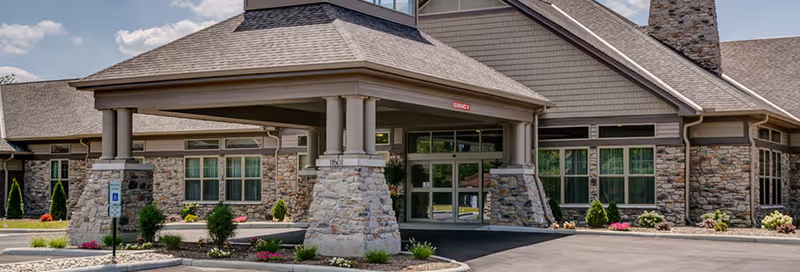 Exterior view of Sanctuary Pointe Nursing & Rehab Center showing the main entrance with a covered drop-off area supported by stone pillars, surrounded by landscaped bushes and flowers under a partly cloudy sky.