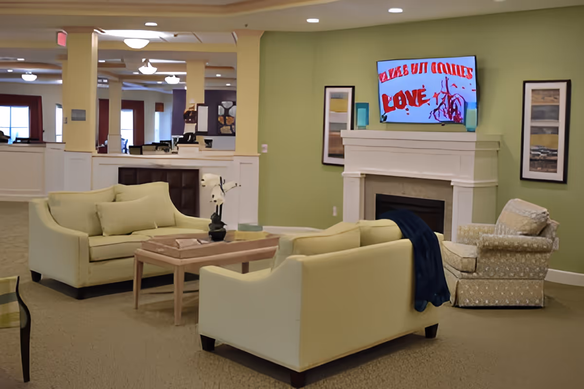A cozy living room area in a senior living facility with two light yellow sofas, a patterned armchair, a wooden coffee table with a small plant and books, a white fireplace with a mounted TV above it, and two framed pictures on a green wall.