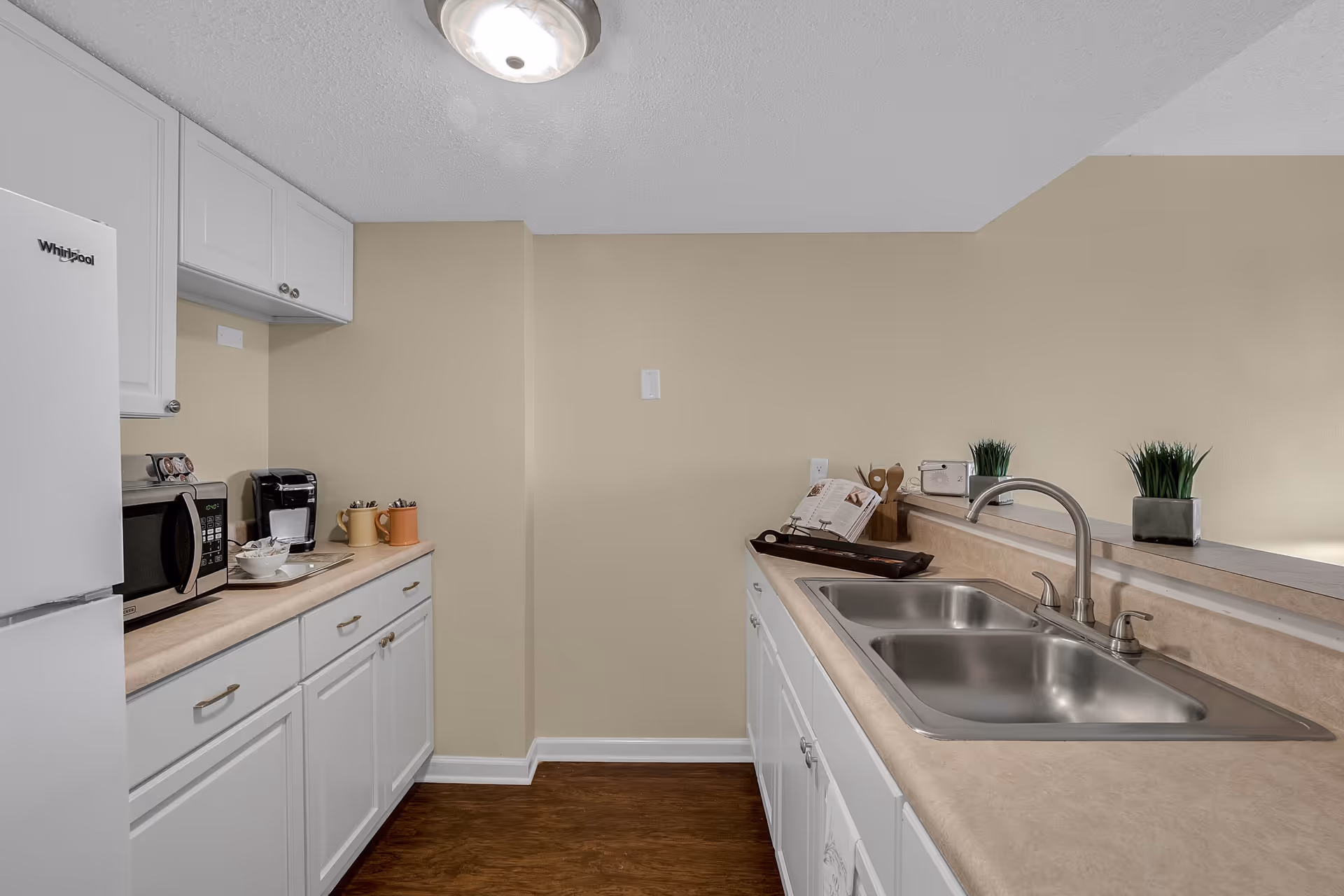 A clean kitchen area with white cabinets and beige countertops. On the left side, there is a white Whirlpool refrigerator, a microwave, a coffee maker, and two mugs. On the right side, there is a double stainless steel sink with a faucet, two small potted plants, a cookbook on a stand, and some kitchen utensils. The walls are painted beige and the floor has a wood finish.