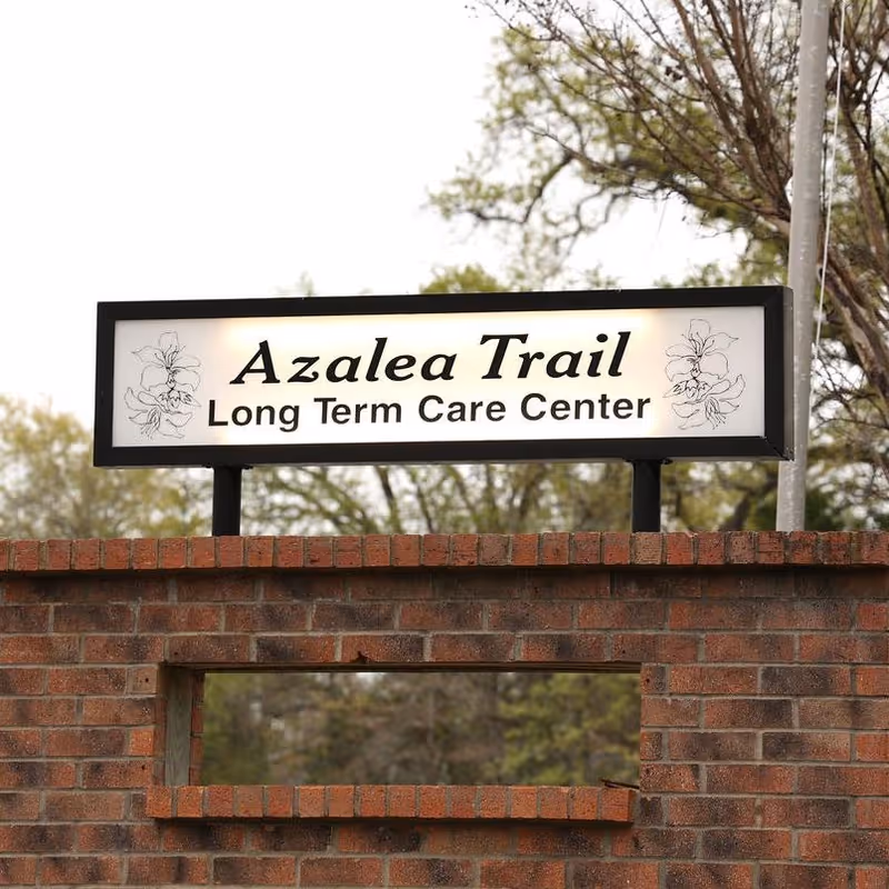 Illuminated sign reading "Azalea Trail Long Term Care Center" mounted above a brick wall with trees in the background.