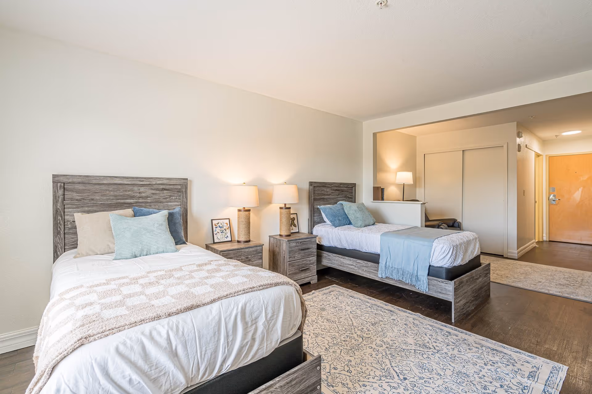 A bright, clean bedroom with two twin beds featuring wooden headboards and white bedding accented with blue and beige pillows. Between the beds is a nightstand with two matching lamps and a framed picture. The room has a patterned rug on the wooden floor and an open doorway leading to a hallway with a wooden door and closet with sliding doors.