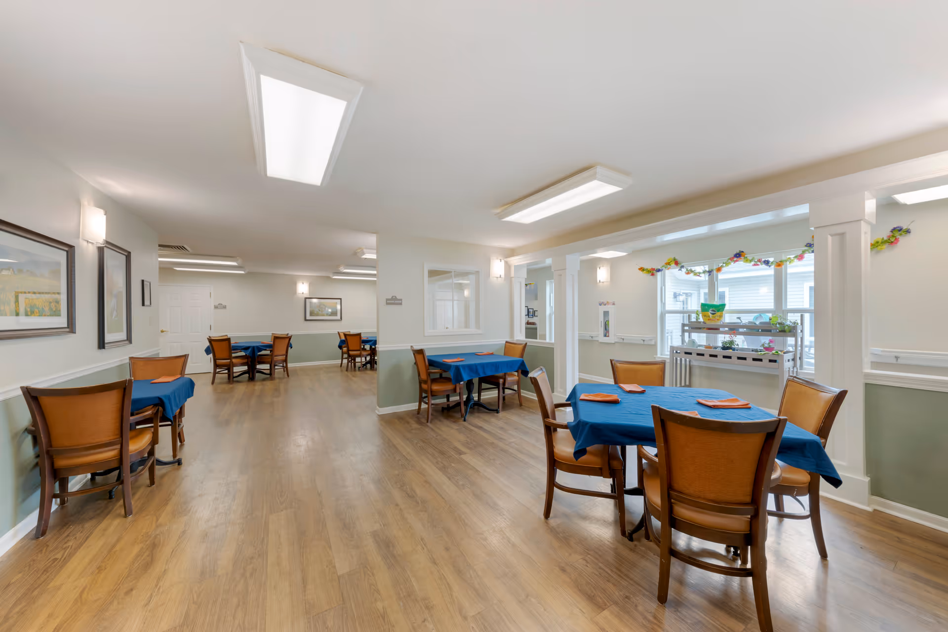 A bright dining room with wooden floors and several tables covered with blue tablecloths and orange napkins. The room has white walls with green wainscoting, framed pictures on the walls, and large windows letting in natural light. There are wooden chairs around the tables and ceiling lights providing additional illumination.