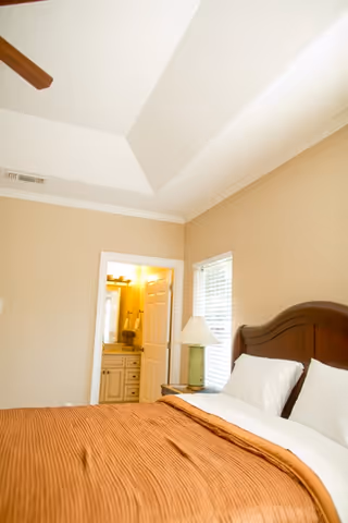A bedroom with a large bed covered in a brown blanket and white pillows. There is a wooden headboard and a bedside table with a lamp. A window with blinds is next to the bed, and an open door leads to a bathroom with a vanity and mirror visible.