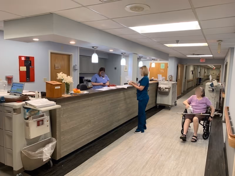 A nursing station in a healthcare facility hallway with two healthcare workers standing behind the counter, one reviewing documents and the other writing. A person in a wheelchair holding a cup is positioned near the counter. The hallway extends into the background with handrails on both sides and overhead fluorescent lighting.