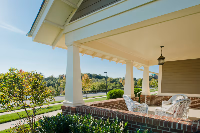 Covered brick porch with white columns and wicker chairs overlooking a landscaped lawn and trees.