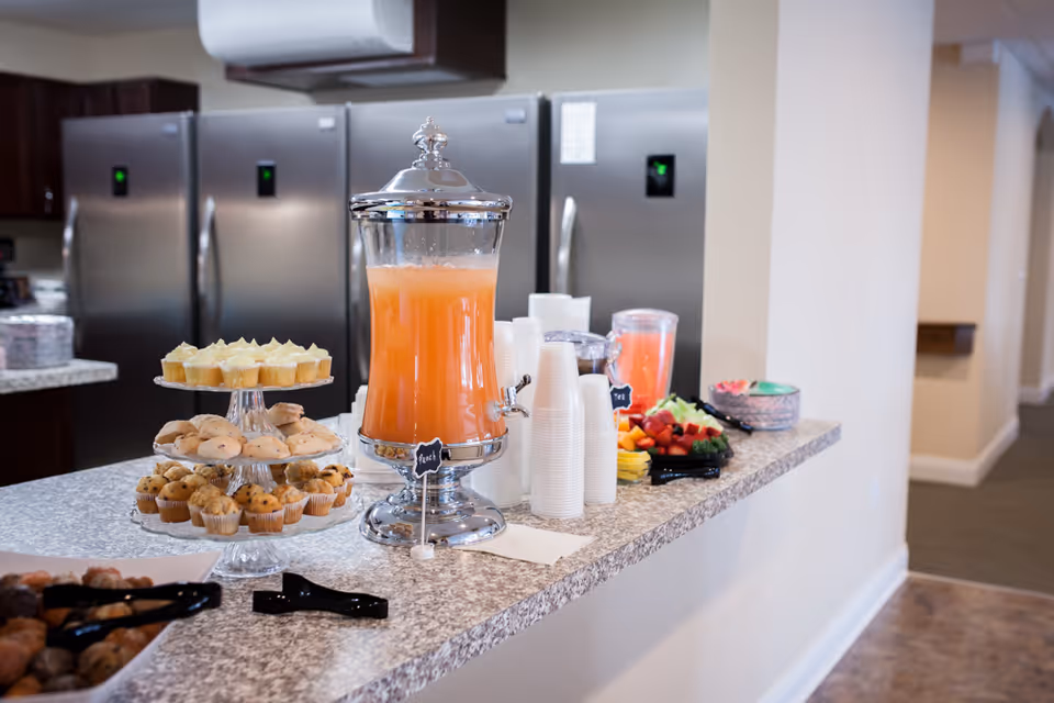 A kitchen counter with a tiered tray of assorted muffins and pastries, a large beverage dispenser filled with orange juice, stacks of white disposable cups, a tray of fresh fruit, and several stainless steel refrigerators in the background.