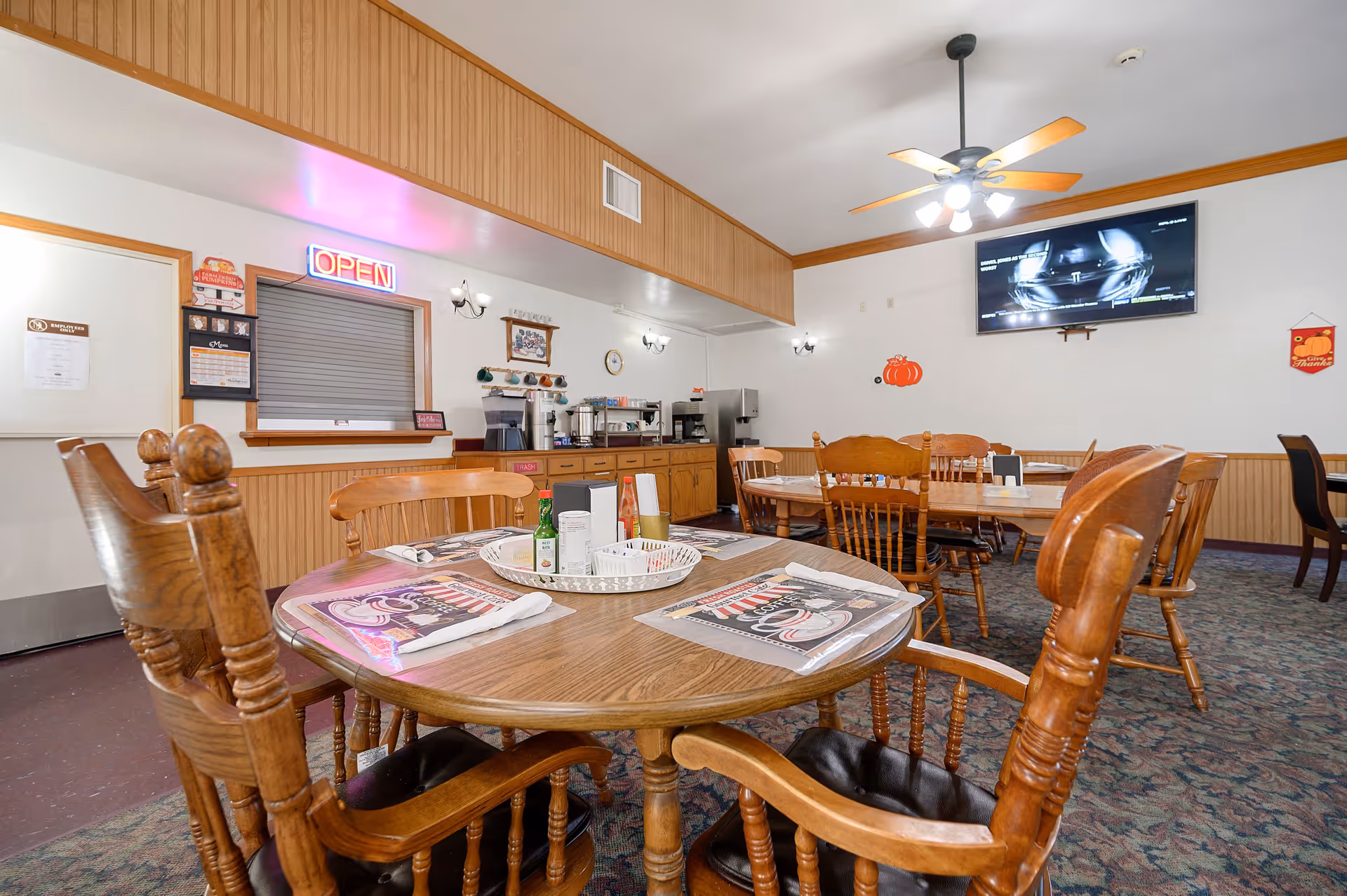 Interior view of a senior living facility dining room with wooden tables and chairs. The tables are set with placemats, napkins, and condiments. There is a serving window with an illuminated 'OPEN' sign above it, and a counter with coffee and beverage machines. A ceiling fan with lights is mounted on the ceiling, and a flat-screen TV is mounted on the wall. The room has wood paneling and carpeted flooring.