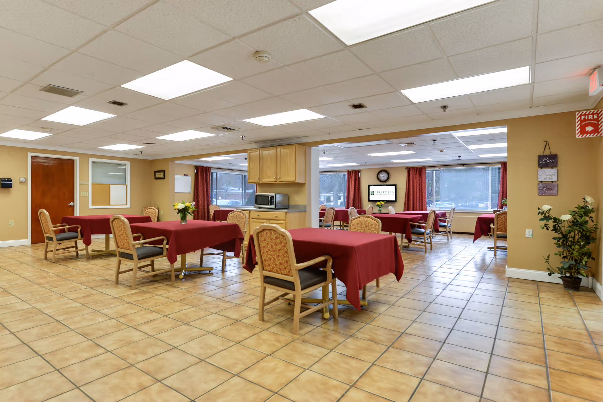 A spacious dining room with several tables covered in red tablecloths and cushioned chairs arranged around them. The room has tiled flooring, beige walls, and a drop ceiling with fluorescent lighting. There are large windows with red curtains letting in natural light, a microwave on a countertop, a clock on the wall, and a TV screen displaying the Crestfield Rehabilitation Center & Fenwood Manor logo. A potted plant and wall decorations are visible on the right side.
