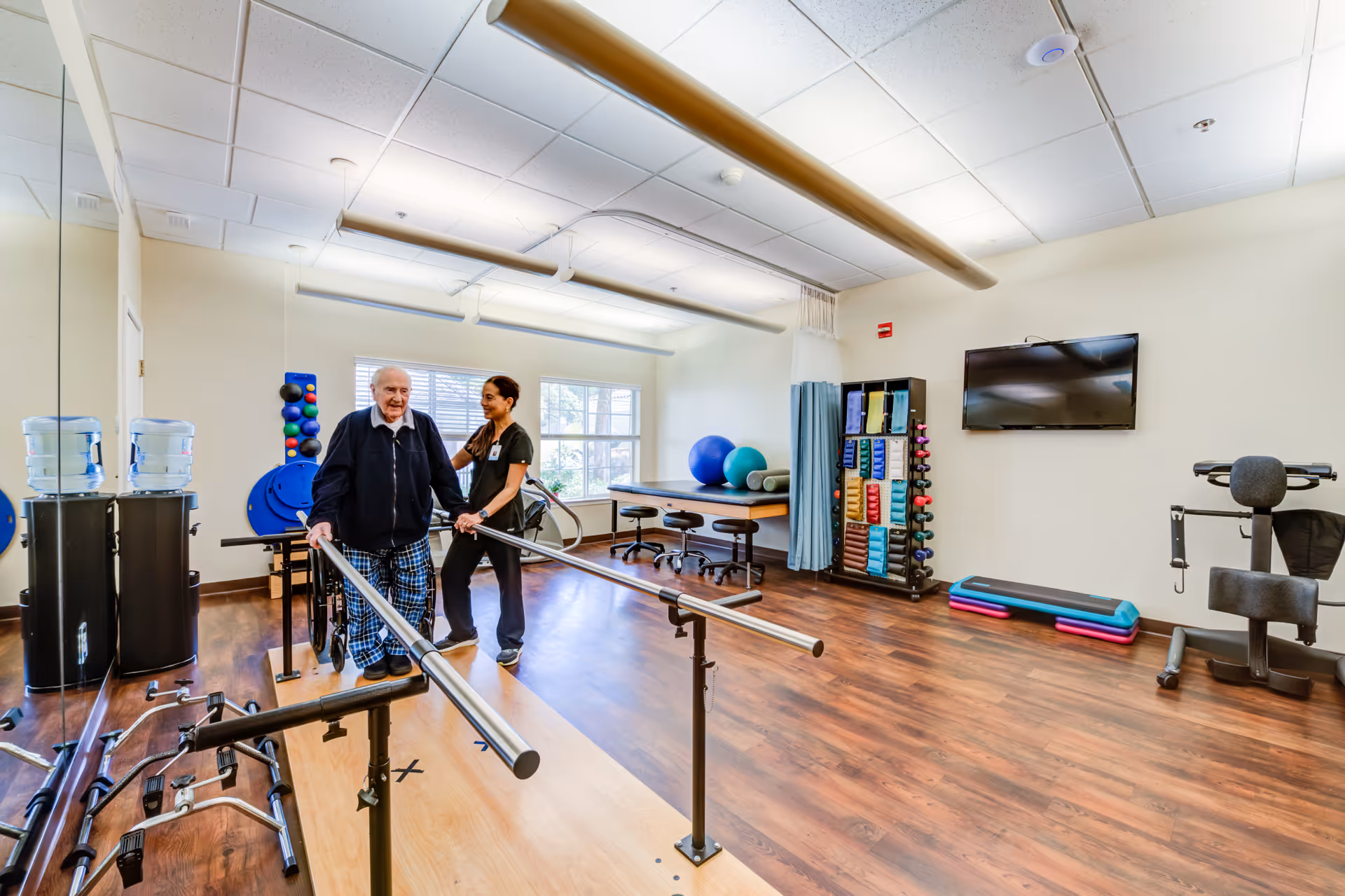 An elderly man is assisted by a healthcare worker while walking between parallel bars in a physical therapy or rehabilitation room. The room has wooden flooring, exercise equipment including weights, exercise balls, and a TV mounted on the wall. There is a large window letting in natural light.