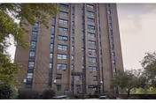 Tall multi-story brick residential building with numerous windows, surrounded by trees and greenery under a partly cloudy sky.